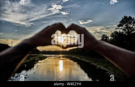 Ricordi estivi concetto. Primi posti per romantiche data. La luce del sole al tramonto atmosfera romantica. Mani maschio a forma di cuore simbolo del gesto di amore e romanticismo. Cuore gesto davanti al tramonto sopra il fiume. Foto Stock