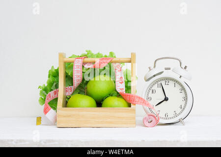 Tempo di sani e diete concetto. Vintage retrò sveglia e mela verde e lattuga in legno cesto su grunge bianco tavola di legno. Wellness e lui Foto Stock