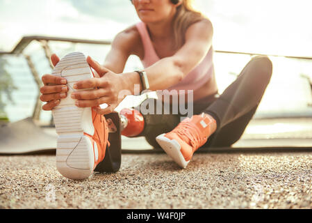 Esercizi di stretching. Foto ritagliata della giovane bella donna di cuffie con protesi di gamba ascoltando musica e stirando la gamba mentre è seduto sul ponte. Sport disabili concetto. La motivazione. Uno stile di vita sano Foto Stock