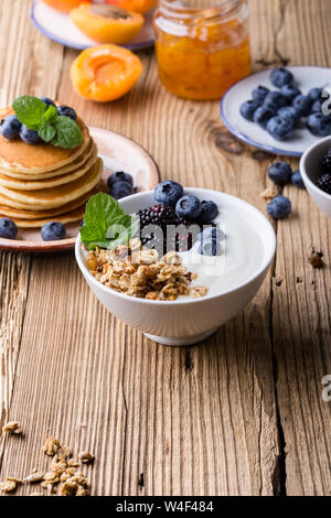 Sana colazione o brunch, preferito pasto mattutino. Muesli fatti in casa con yogurt, frittelle, fresca estate bacche e frutta, marmellata sul tavolo di legno Foto Stock
