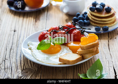 Una sana colazione vegetariana o il brunch, cibo preferito. Uova fritte con pomodorini, frittelle, fresca estate frutti, bacche, caffè e succo di frutta, a Foto Stock