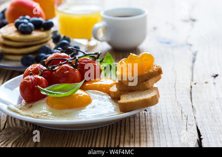 Una sana colazione vegetariana o il brunch, cibo preferito. Uova fritte con pomodorini, frittelle, fresca estate frutti, bacche, caffè e succo di frutta, a Foto Stock