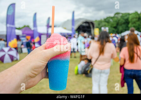 Un maschio di mano che tiene un slushie bere in un campo in un festival. Un slushie è una bevanda fatta dal ghiaccio aromatizzato. Foto Stock