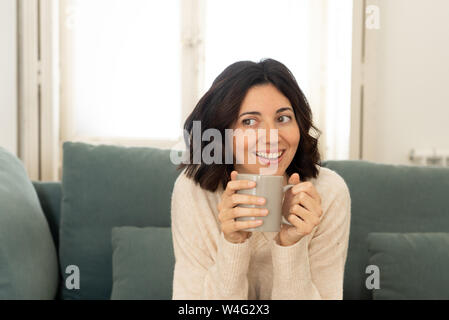 Stile di vita Ritratto di giovane abbastanza rilassato donna bere il caffè caldo al cioccolato o sentirsi felice e accogliente a casa sorridendo felice sul lettino. In leisur Foto Stock
