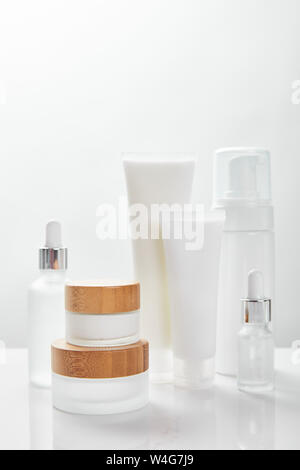 Superficie bianca con le bottiglie di vetro, crema nei tubi, vasi ed erogatore cosmetico Foto Stock