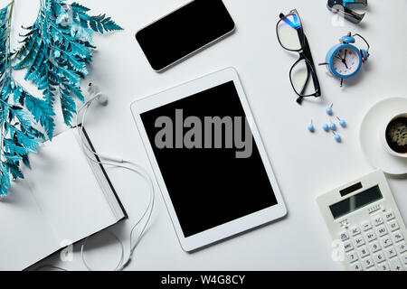 Vista superiore della tavoletta digitale, smartphone, notepad, auricolari, caffè, cancelleria, orologio con sveglia, calcolatrice, bicchieri e ramo blu sulla superficie bianca Foto Stock