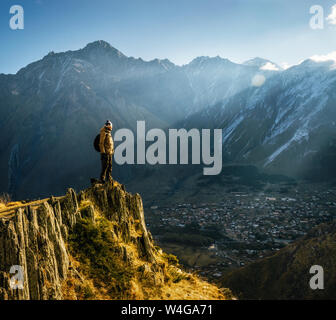 Giovani turisti in bright hat, pantaloni neri con uno zaino sorge su cliff's edge e guardando il misty Mountain Village e il ghiacciaio al sunrise Foto Stock