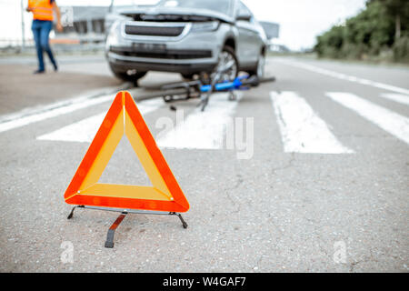 Scena di un incidente stradale con auto e biciclette rotte giacente sul passaggio pedonale. Triangolo di traffico in primo piano Foto Stock