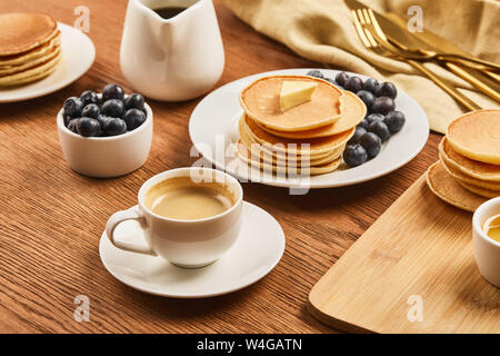 Una gustosa prima colazione con tazza di caffè, frittelle e mirtilli vicino al telo di lino con posate su una superficie di legno Foto Stock