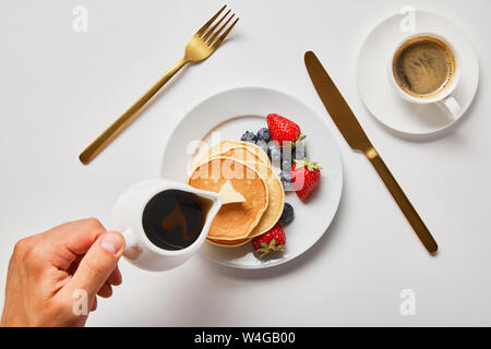 Vista ritagliata dell uomo di sciroppo di colata sulle frittelle con frutti di bosco vicino a golden posate e tazza di caffè Foto Stock