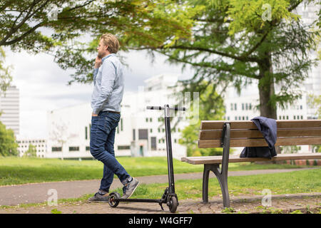 Imprenditore con E-scooter sul telefono nel parco cittadino, Essen, Germania Foto Stock