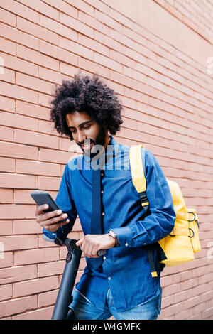 Ritratto di sorridente giovane con zaino e E-Scooter guardando il telefono cellulare di fronte a un muro di mattoni Foto Stock