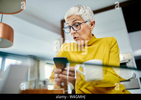 Donna senior di bere caffè, controllo messaggi di testo sullo smartphone Foto Stock