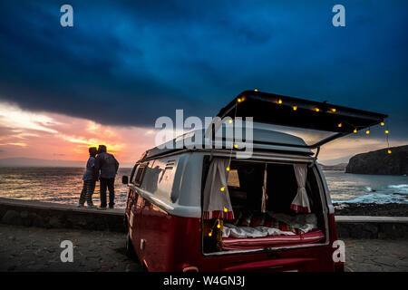 Coppia senior in viaggio in un'annata van, kissing al tramonto al mare Foto Stock