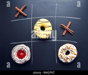 Ciambelle vetrato tic-tac-toe gioco. Chalk disegno su uno sfondo scuro. Cucina creativa laici piana. Gioco una partita di tris con ciambelle sulla lavagna. Unheal Foto Stock