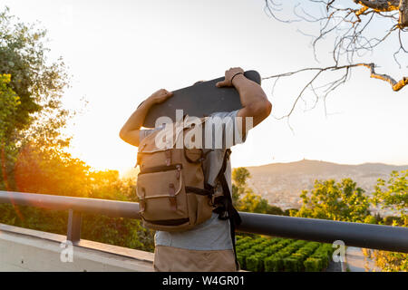 Vista posteriore del giovane con lo skateboard sulle sue spalle al tramonto Foto Stock