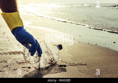 La mano di un uomo con i guanti di prelevare la bottiglia di plastica che inquinano la spiaggia, pulizia sulla riva, campagna per pulire il concetto di volontariato Foto Stock