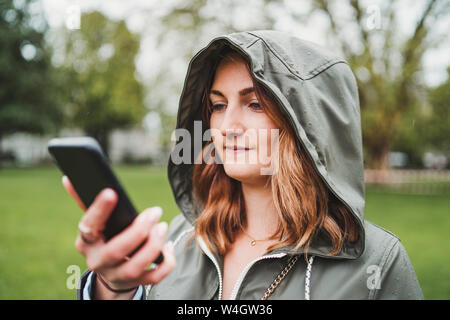 Giovane donna indossando giacca con cappuccio e utilizzando il telefono cellulare in un giorno di pioggia Foto Stock