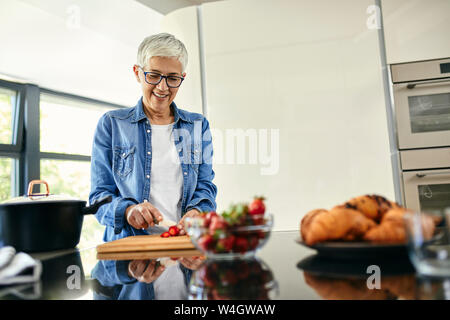 Senior woman standing in cucina, tritare le fragole Foto Stock