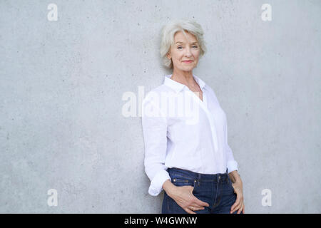 Ritratto di rilassata donna matura che indossa una camicia bianca appoggiata contro la parete di cemento Foto Stock