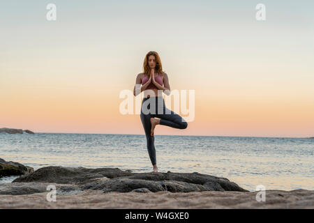 Giovane donna a praticare yoga sulla spiaggia, facendo la posizione dell'albero, durante il tramonto in spiaggia tranquilla, Costa Brava, Spagna Foto Stock