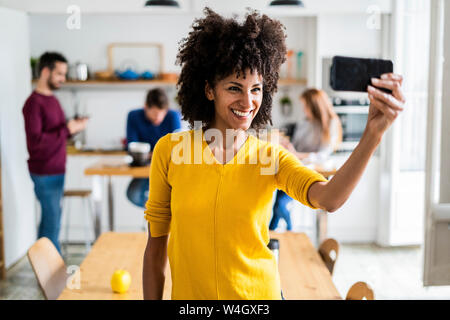 Donna felice di prendere una selfie a casa con gli amici in background Foto Stock