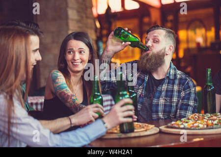 Amici avente un drink in un bar, essi sono seduti ad un tavolo di legno con birre e pizza Foto Stock