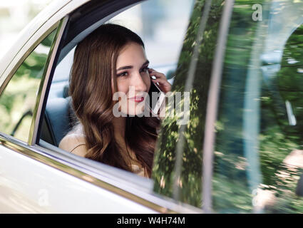 Una giovane ragazza è seduta sul sedile posteriore di un auto e sta parlando al telefono. Foto Stock