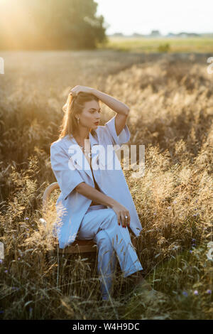 Foto di moda di attraente donna bionda in blu tuta moda seduti su una sedia in campo di grano. Foto Stock