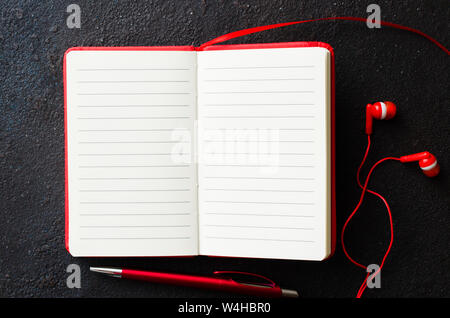 Vuoto notebook rosso con penna rossa e cuffie su sfondo scuro. Carta bianca per il testo. Appartamento laico, vista dall'alto, spazio di copia Foto Stock