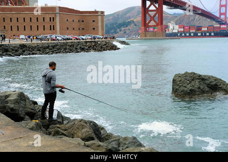 Giovane americano africano pesca maschio mentre un Yang Ming marine nave da trasporto vele sotto il Golden Gate Bridge di San Francisco, California. Foto Stock