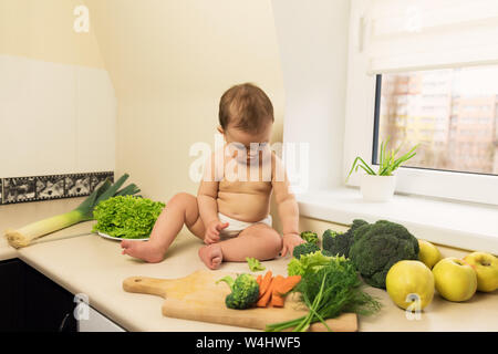 Il bambino nel pannolino è seduta sul tavolo della cucina. Un bambino gioca e si diverte con freschi ortaggi e frutta. Foto Stock