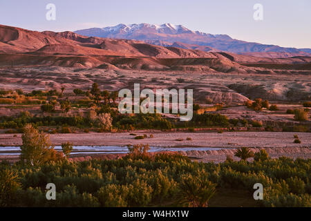 Il fiume scorre attraverso il deserto roccioso in Marocco Africa con coperta di neve montagne Atlas in background Foto Stock