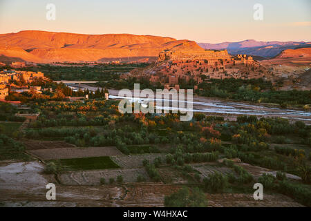 La mattina presto Sole, colpendo la parte superiore del patrimonio storico di Ait Ben Haddou con oasi di verde nel deserto circondato da montagne aride in Marocco Foto Stock