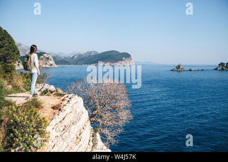 Un turista con lo zaino sulla cima di una scogliera o collina vicino al mare appare in lontananza. Viaggiare da soli. Foto Stock