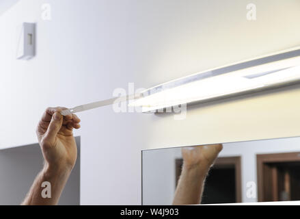 Installare la lampada sopra lo specchio. Un uomo è la rimozione di un nastro protettivo dalla lampada dopo l'installazione. Foto Stock