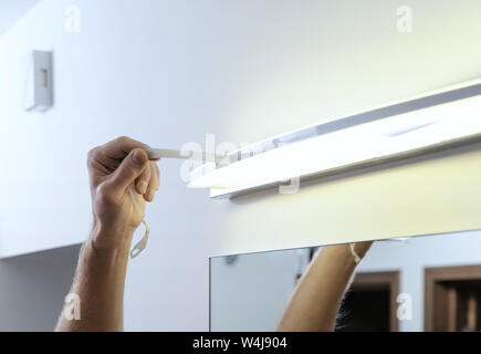 Installare la lampada sopra lo specchio. Un uomo è la rimozione di un nastro protettivo dalla lampada dopo l'installazione. Foto Stock