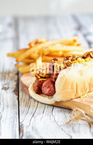 Hot Dog con chili, il formaggio cheddar e senape. Messa a fuoco selettiva con sfocato patatine fritte in background. Foto Stock