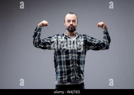 Macho uomo barbuto in casual maglietta a maniche lunghe in piedi flettendo le braccia con un wry espressione in un tentativo di mostrare il suo bicipite isolato su grigio Foto Stock