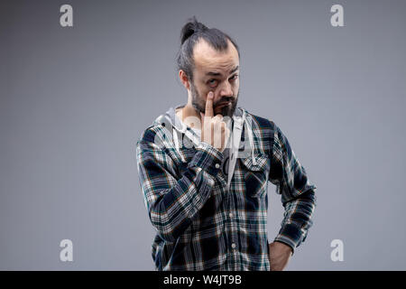 Uomo Barbuto con coda di cavallo tirando verso il basso una palpebra con il suo dito in un gesto di avvertimento per essere desti e vigilanti isolato sul grigio con spazio di copia Foto Stock