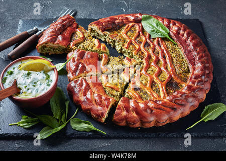 Close-up di fettine di spinaci Torta di ricotta su un nero ardesia vassoio con salsa tzatziki in una ciotola su una tavola di cemento, orizzontale vista da sopra Foto Stock