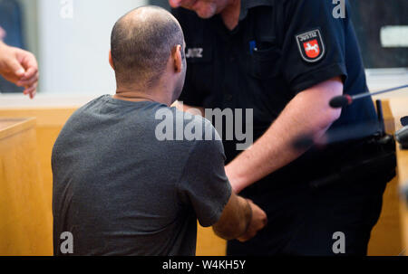 24 luglio 2019, Bassa Sassonia, Hannover: Il convenuto è seduta in Aula di Hannover Corte Regionale quando un funzionario del tribunale rimuove il suo manette. Secondo l'accusa, l'uomo, nato nel 1983, distribuiti e acceso il fuoco acceleratori in un edificio di appartamenti in Hanover-Ahlem nel febbraio 2019. Foto: Julian Stratenschulte/dpa Foto Stock