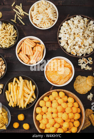 Tutti i classici snack di patate con arachidi, popcorn e anelli di cipolla e salati pretzel nella ciotola piastre su legno. Ghirigori con bastoni e patatine e cr Foto Stock