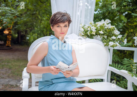 Giardino estivo, buon umore, stile retrò. adulti sognanti brunette di cinquanta seduto su un romantico bianco banco vintage con un libro nelle sue mani. ritratto di Foto Stock