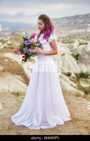 Donna con un bel bouquet di fiori nelle sue mani si erge sulla montagna in raggi dell'alba al tramonto. Bella bianca lunga veste sulla ragazza b Foto Stock