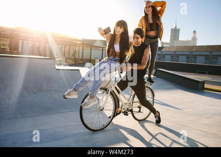 Donna seduta sul manubrio di bici tenendo selfie con gli amici. Giovane donna prendendo un autoritratto con i suoi amici in bicicletta. Foto Stock