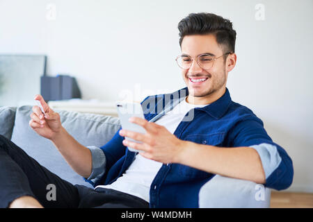 Immagine del giovane uomo seduto sul divano di casa e pagare per gli acquisti online con lo smartphone e carta di credito Foto Stock