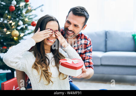 Allegro uomo cercando di sorpresa fidanzata con il regalo di Natale. La famiglia durante la celebrazione Foto Stock