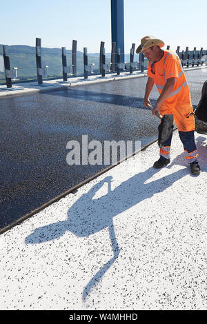 Rachtig Zeltingen, Germania. Il 24 luglio, 2019. Lavoratori asfalto la superficie stradale del ponte Hochmosel in costruzione a temperature ben al di sopra di 30 gradi Celsius. Non vi è alcun luogo ombroso su 160 metri di un edificio alto sopra la valle di Mosel. Credito: Thomas Frey/dpa/Alamy Live News Foto Stock