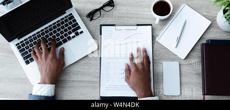 Irriconoscibile afro imprenditore lavorando sul suo computer portatile con i report finanziari intorno Foto Stock